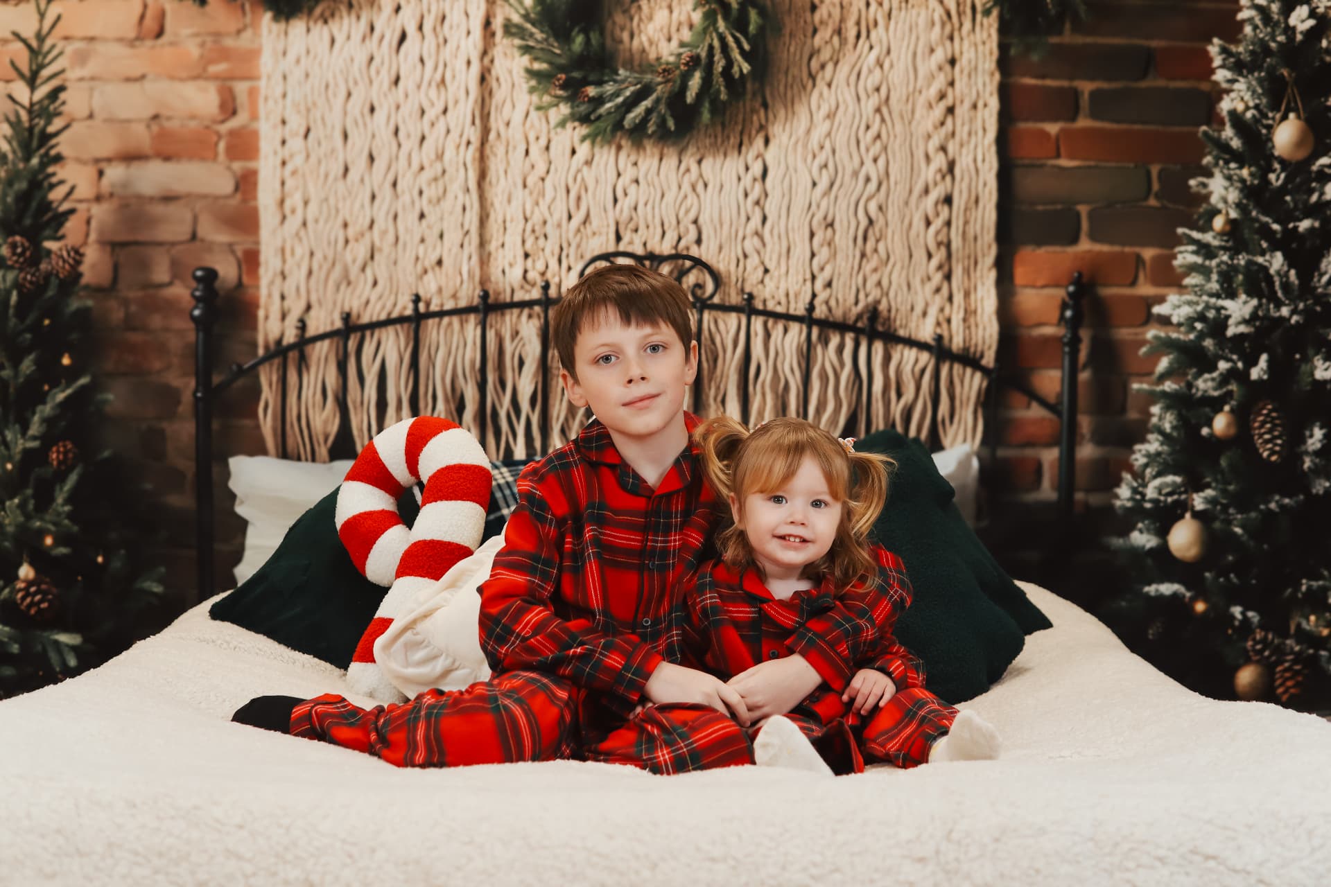 Two children in matching red plaid pyjamas sitting on a cosy bed with Christmas decor