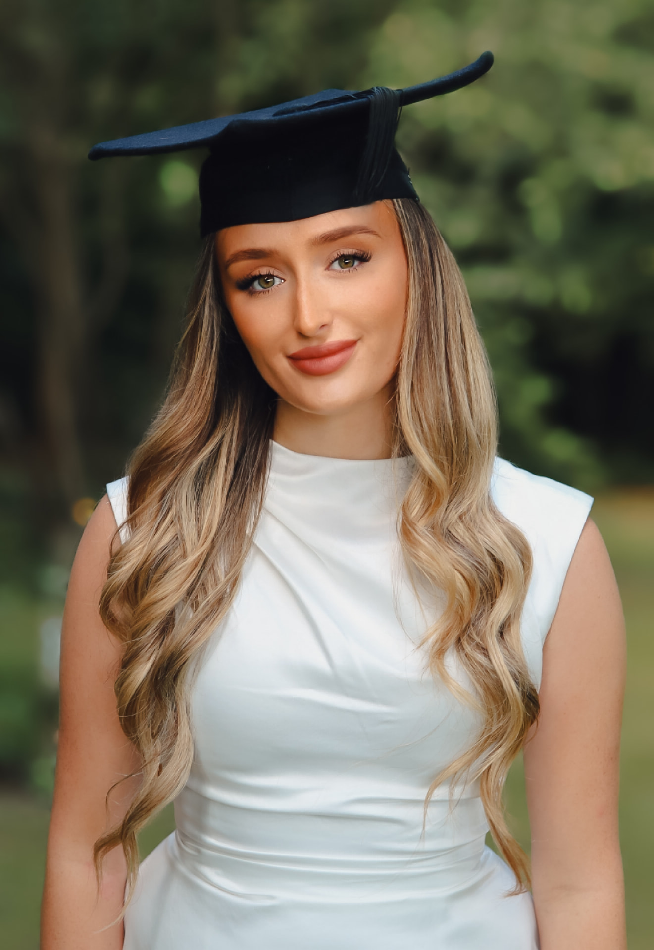 Graduation portrait of a young woman in a cap and gown in dappled garden light