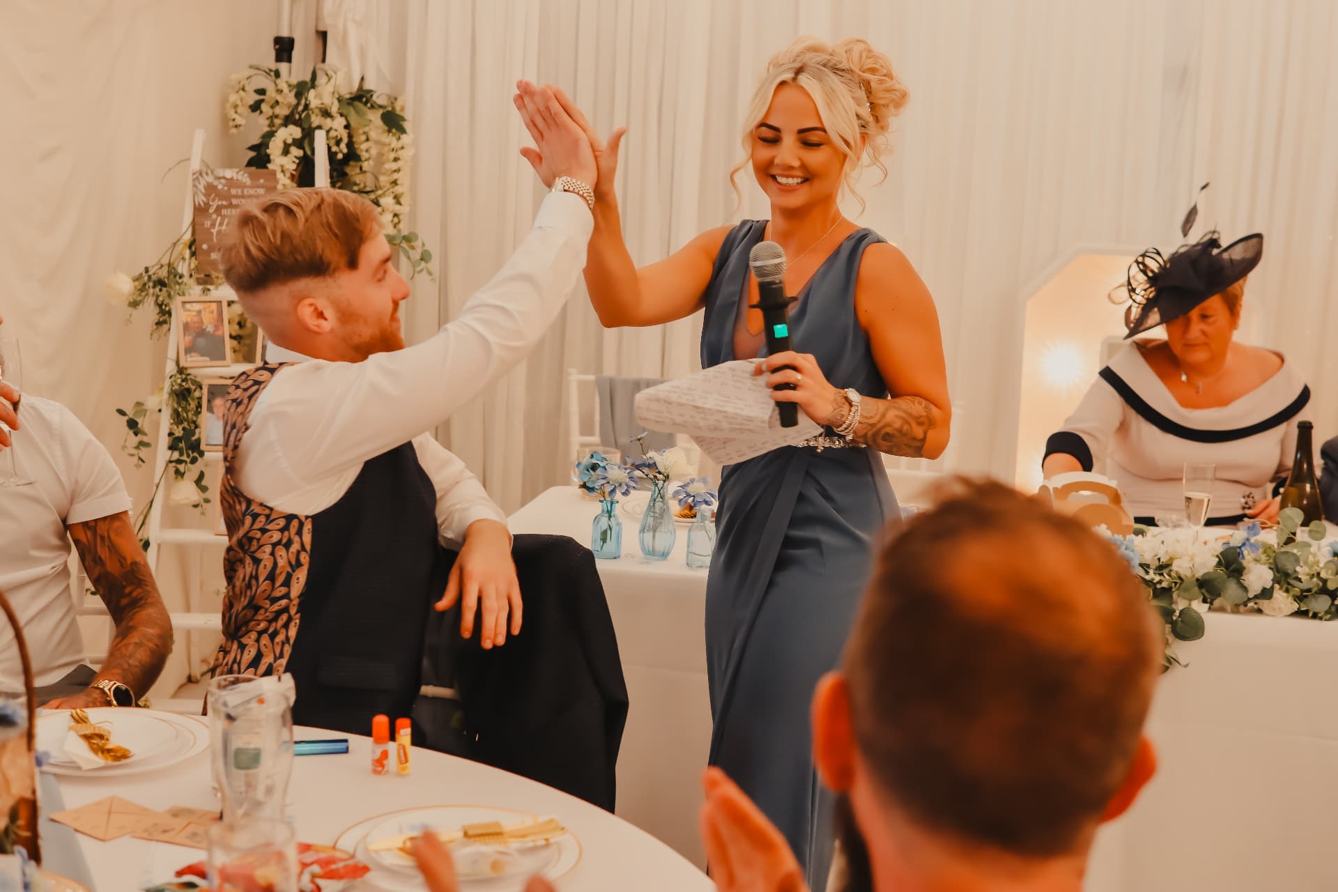 Wedding guest in a blue dress holding a microphone and gift gives a joyful high-five to a seated guest during the reception speeches