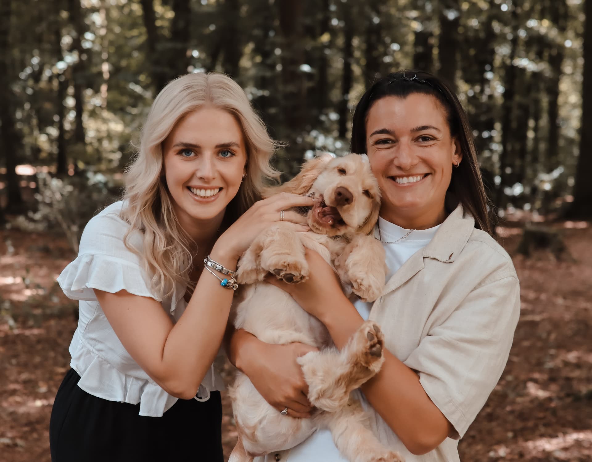 Two women smiling while holding a happy golden retriever puppy in a sunlit woodland