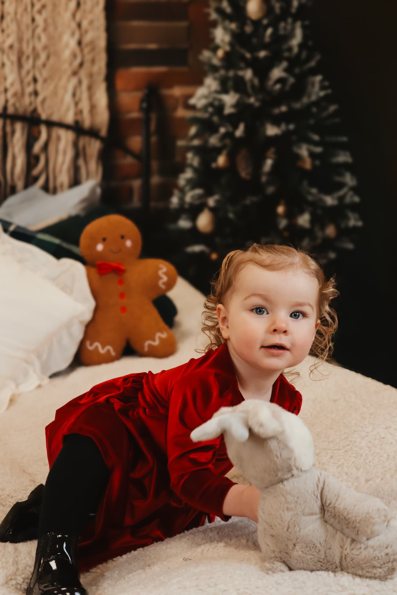 Toddler in a red velvet dress reaching for a soft toy beside a Christmas tree