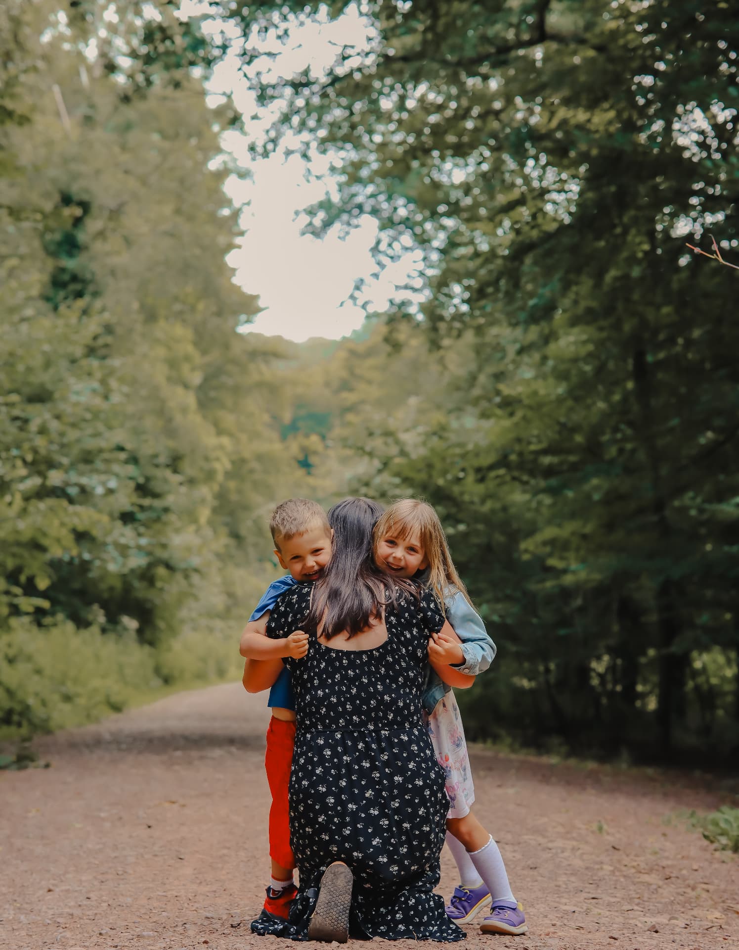 A mother kneeling on a woodland path while two young children hug her from either side